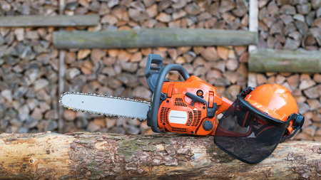 Portable gasoline chainsaw and helmet with protective mesh visor on a wooden log. Professional...