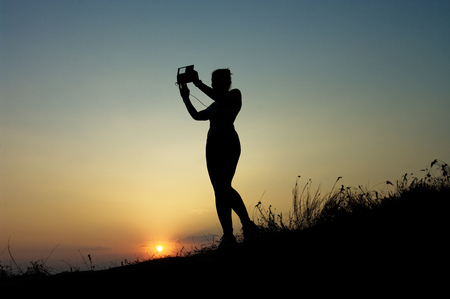 Silhouette of a young woman doing selfi at sunset sky backgroundの写真素材