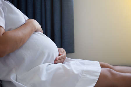 Close-up of Asian pregnant woman touching her belly on the foreground on her bed at homeの写真素材