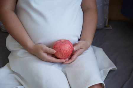 Cropped image of Asian pregnant woman sitting on bed with red apple in her hands