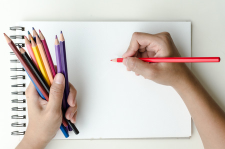 Man holding a fistful of colored pencils in one hand while commencing sketching in a sketch book to show off his creativity and artistic skills with the other, view from aboveの写真素材