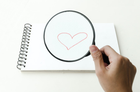 Hand-drawn red romantic heart drawn on a blank page in a sketch book and viewed through a magnifying glass held in the hand of a man, conceptual of love and romanceの写真素材