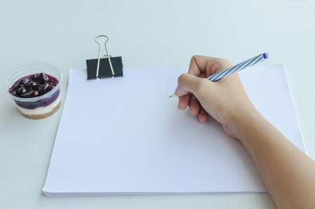 Close-up of a woman writing into blank paper with binder clip beside cup of blueberry chesse pineの写真素材