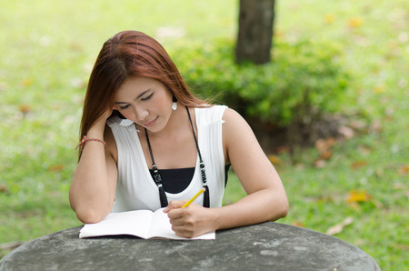 Beautiful young redhead Asian woman writing in a notebook while sitting outdoors in a park smiling and looking to the cameraの写真素材