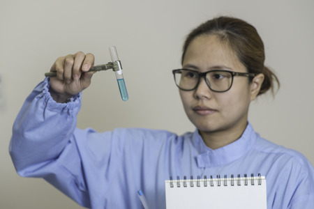 Young female Asian lab worker wearing spectacles reading a chemical test checking the color of the solution in a test tubeの写真素材