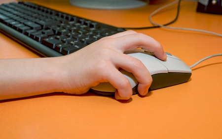Children's right hand on the mouse on the background of computer keyboard on the orange tableの写真素材