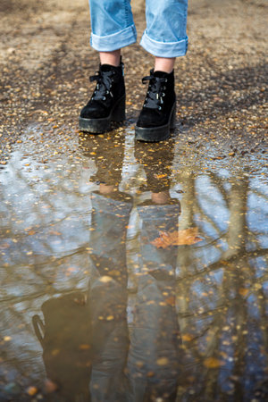 Bottom half of a person's legs wearing boots and standing in front of a puddle of mud in a forest with the person and the trees reflecting in the water during autumnの写真素材