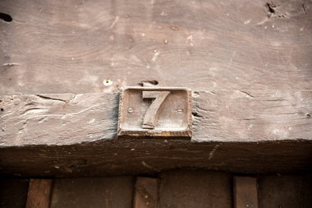 Rustic metal house number seven mounted on a weathered wooden beam, showing patina and worn paint, a...