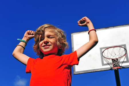 Happy smiling boy celebrating victory with his hands up with basketball hoop on backgroundの写真素材