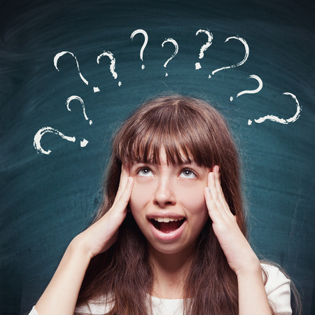 Young girl with questioning expression and question marks above her head and blackboard in the backgroundの写真素材