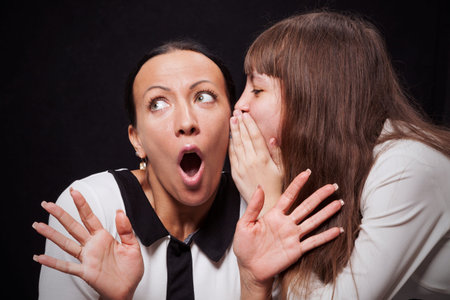 Girl speaks ear of a young surprised woman on a black background