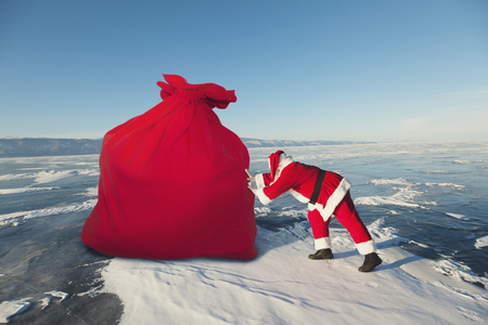 Santa Claus pulls big red bag on the frozen lake in winterの写真素材