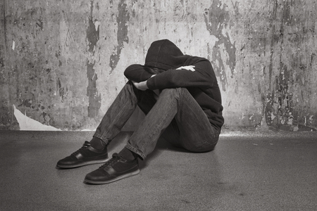 Boy teenager sitting with his head down near the tattered walls in the roomの写真素材