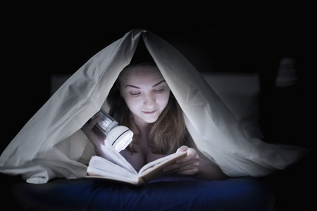 Teenage girl reading a book in bed under the covers of her flashlight lightingの写真素材