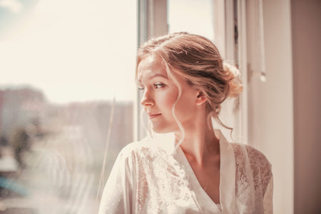 Young beautiful girl in a white peignoir and wedding veil looks out the window
