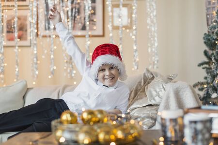 New Year mood: Ready for the New Year! The boy smiles, lying on a sofa in a Santa Claus hat, in a New Year's decorated roomの写真素材