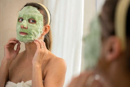 Adult beautiful woman in a bathroom and a towel takes care of her skin applies a green tissue wellness face maskの写真素材