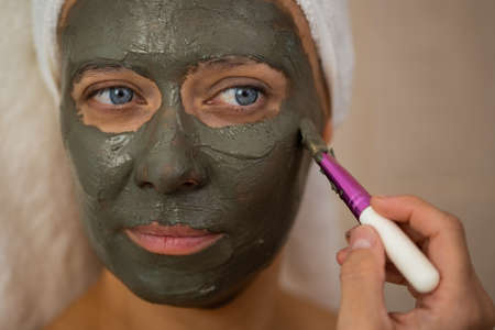 An adult beautiful woman in a bathroom and a towel takes care of her skin and applies a clay mask to her faceの写真素材