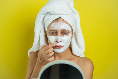 Beautiful woman wrapped in a towel looks in the mirror and applies healing cream to her face on a...