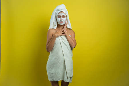 Beautiful woman wrapped in a towel with anti-wrinkle skin cream puts on a mask on her face on a yellow backgroundの写真素材