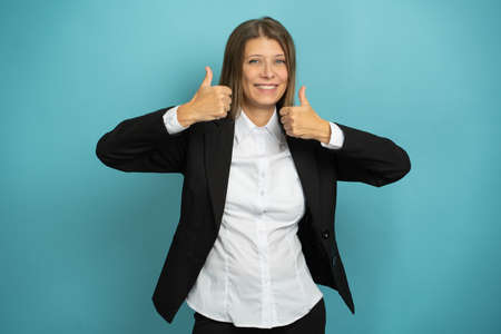 Beautiful Adult Business Woman In Black Suit And White Shirt Shows Thumb Up On Blue Backgroundの写真素材