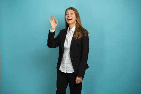 Beautiful Adult Business Woman In Black Suit And White Shirt Shows Fingers OK On Blue Backgroundの写真素材