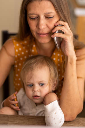 A beautiful woman at home in the kitchen uses a naotbook and tries to work with a small child in her arms and communicate on the phoneの写真素材