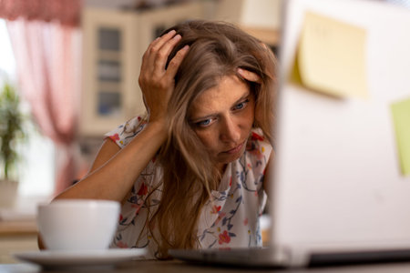 Tired emotional and expressive beautiful adult woman at home in the kitchen working on a laptop