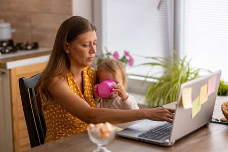 Beautiful woman at home in the kitchen uses naotbook and tries to work with a small child in her armsの写真素材