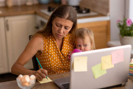 Beautiful woman at home in the kitchen uses naotbook and tries to work with a small child in her armsの写真素材