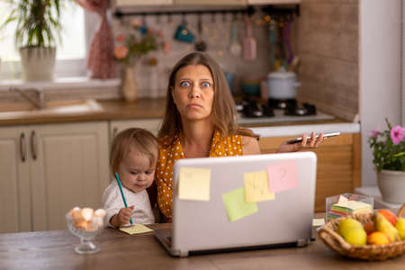 Beautiful woman at home in the kitchen uses naotbook and tries to work with a small child in her armsの写真素材