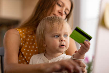 A beautiful woman at home in the kitchen uses a laptop and tries to work with a small child in her arms and buy purchases online with a credit cardの写真素材