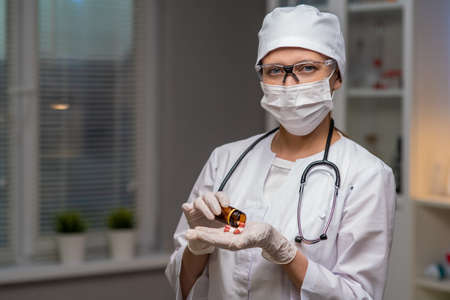 Female Doctor In Laboratory Cabinet On Background Of Test Tubes And Medical Equipment Masked Gloves And Glasses Holds Pills In Handsの写真素材