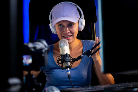 Beautiful Woman In A Cap And White Headphones Leads A Live Stream Through The Internet. Speaks Into Microphone. Live Blogger Talking To Camera On Computer With Monitorsの写真素材