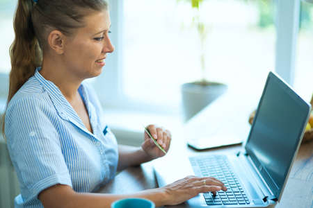Woman housewife at home in the kitchen sits at a table with a laptop. Communicates, relaxes, works from home and online shoppingの写真素材