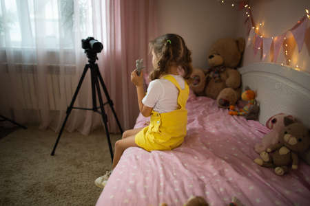 A little girl has her own video blog. She talks about her toys on a digital video camera. Communicates live stream with her followers. Shows them sitting in her room at home on the bed.の写真素材