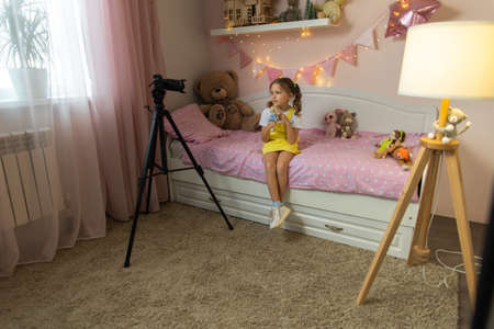 A little girl has her own video blog. She talks about her toys on a digital video camera. Communicates live stream with her followers. Shows them sitting in her room at home on the bed.の写真素材