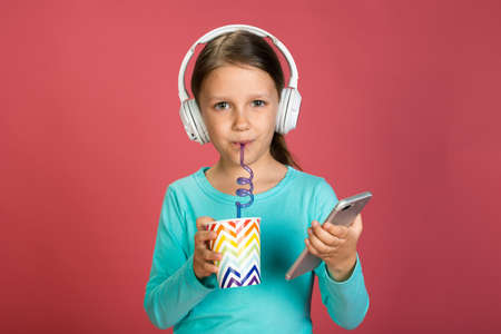 Little Beautiful Baby Girl Pink Background Bright Clothes Yellow Pants Turquoise Blue Shirt Wearing White Headphones Listening To Music With Smartphone In Handの写真素材