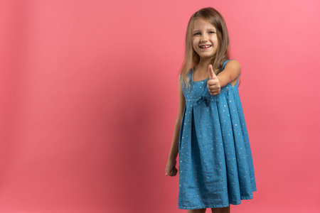 Little beautiful child girl in blue dress showing thumb up on pink background