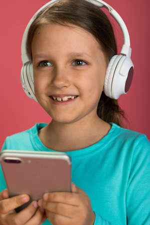 Little Beautiful Baby Girl Pink Background Bright Clothes Yellow Pants Turquoise Blue Shirt Wearing White Headphones Listening To Music With Smartphone In Handの写真素材
