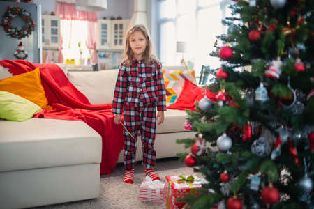 Little Girl In Pajamas Decorates Christmas Tree With Toys And Gift At Home In Roomの写真素材