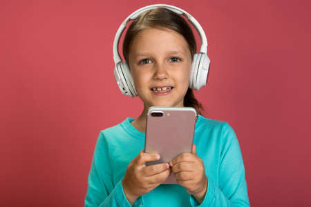 Little Beautiful Baby Girl Pink Background Bright Clothes Yellow Pants Turquoise Blue Shirt Wearing White Headphones Listening To Music With Smartphone In Handの写真素材