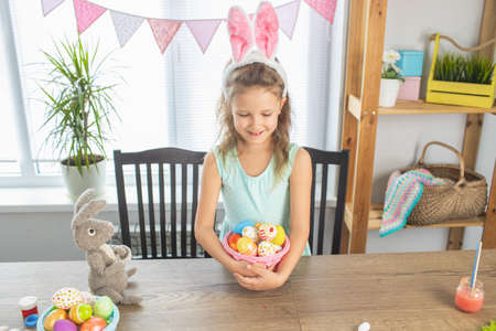 A preschool girl paints eggs for the bright holiday of Easter. Happy at the table with paints and eggsの写真素材
