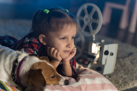 Girl child watching an old movie on a retro vintage film projector with a dog Jack Russell Terrier at homeの写真素材
