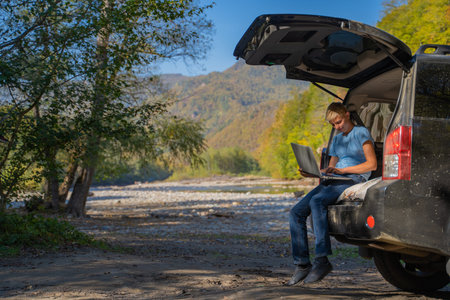 Young man resting in nature with an off-road vehicle resting in the trunk of a car. Uses a laptop...
