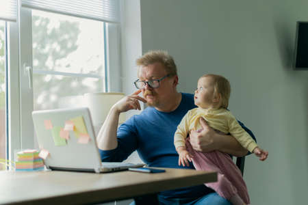 A freelance man father tries to work at home on a computer on the Internet and sits with a small child who prevents him from doing businessの写真素材