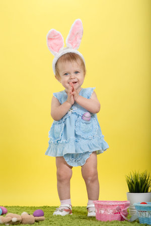 Little baby girl on green grass and yellow background in the ears of an easter bunny with painted...