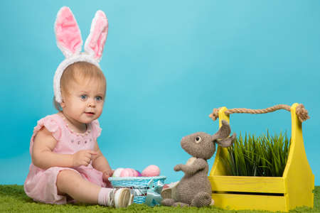 Little baby girl on green grass and blue background in the ears of an easter bunny with painted easter eggs preparing for easterの写真素材
