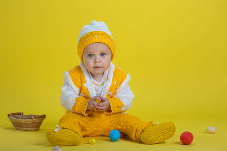 Little child one year age girl in yellow clothes with easter eggs sits on a yellow background expresses emotionsの写真素材
