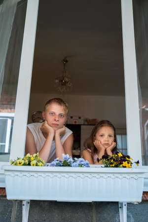 Brother boy teenager with sister girl preschooler look out the window of his home during isolation from the coronavirus pandemicの写真素材